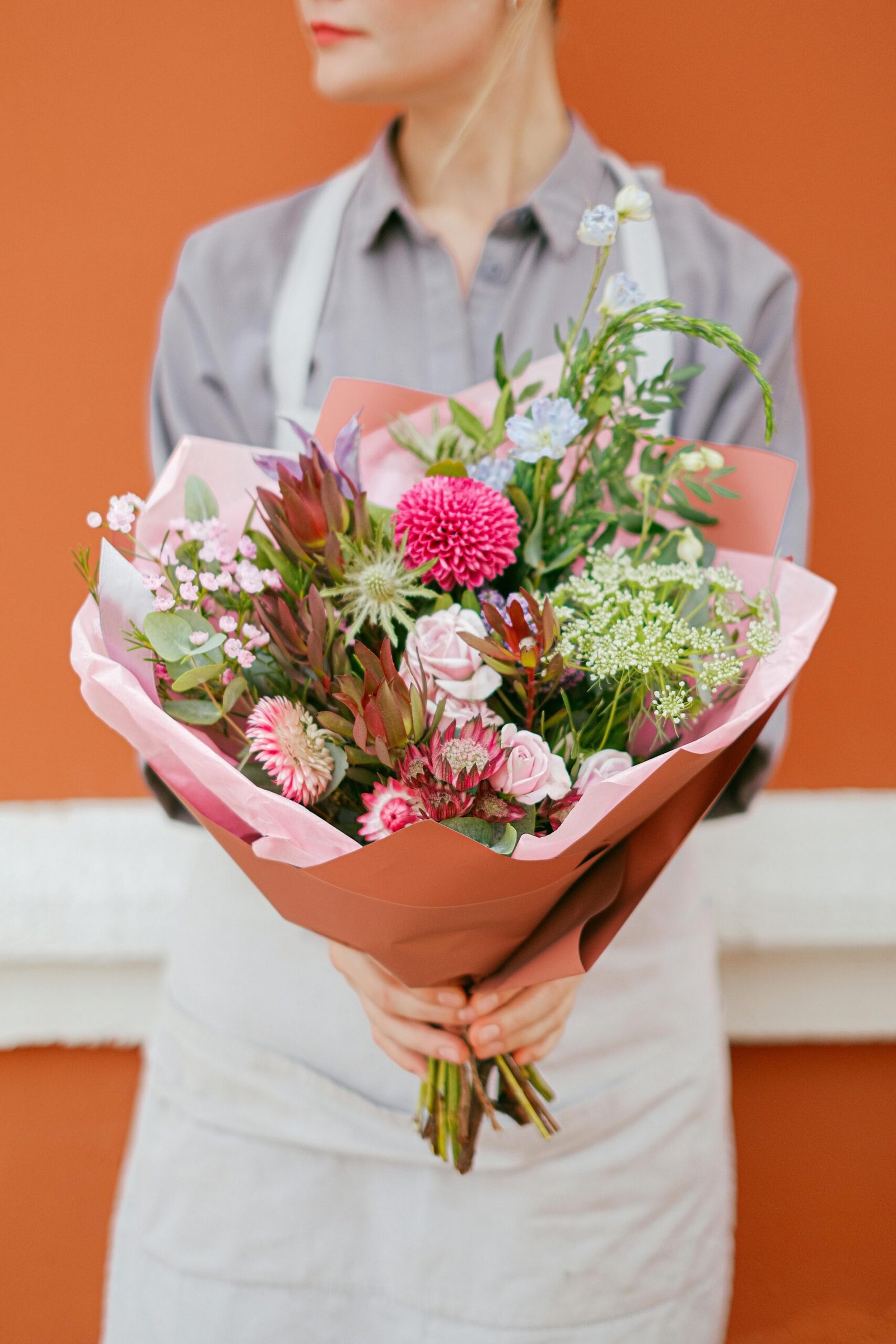 Ramo de flores variadas en tonos rosados y verdes, envuelto en papel elegante y sostenido por una florista frente a fondo naranja.